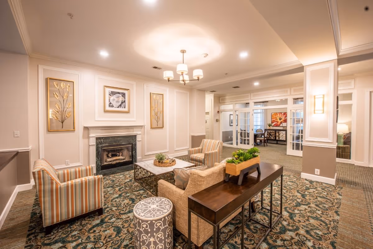 A cozy living room area with a fireplace, two striped armchairs, a beige sofa, a decorative round stool, and a wooden console table with a planter. The room features patterned carpet, framed artwork on the walls, recessed lighting, and a chandelier. In the background, there are glass double doors leading to another room with a table and chairs.