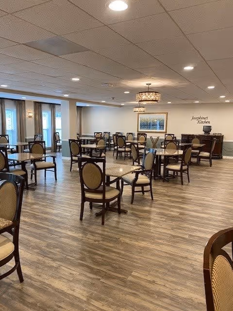 A spacious dining room with multiple tables and chairs arranged neatly on a wood-patterned floor. The room has a drop ceiling with recessed lighting and two decorative hanging light fixtures. Large windows with curtains line one side of the room, and there is a framed picture and a sign on the far wall that reads 'Independent Kitchen'.