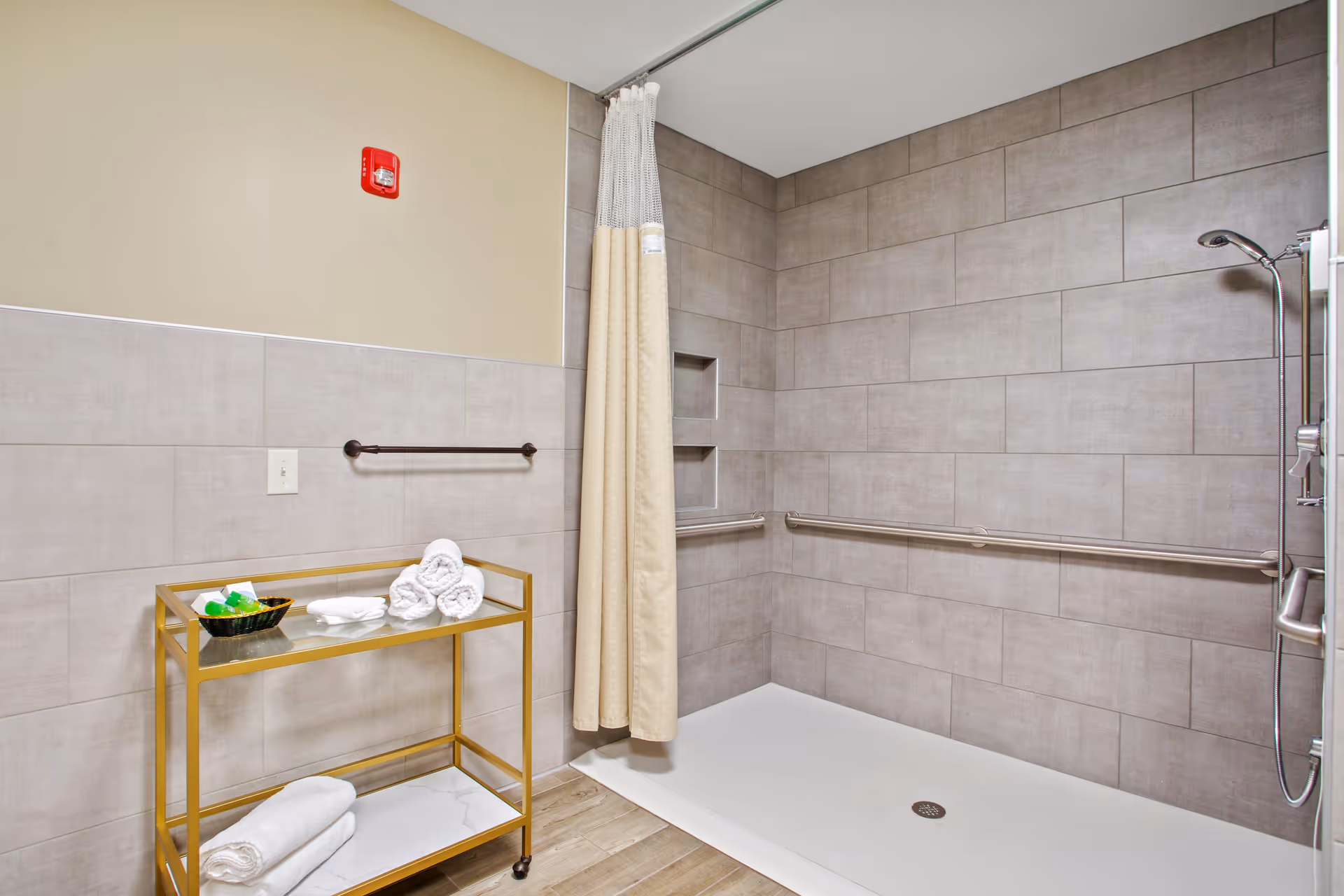 An accessible shower featuring grab bars, a shower curtain, and a gold cart with towels and toiletries.