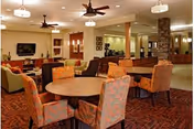 A spacious and warmly decorated common area in a senior living facility featuring multiple round tables surrounded by orange patterned chairs. The room includes ceiling fans, pendant lights, a large flat-screen TV mounted on the wall, and a stone pillar. The carpet has a geometric pattern, and the overall ambiance is cozy and inviting.