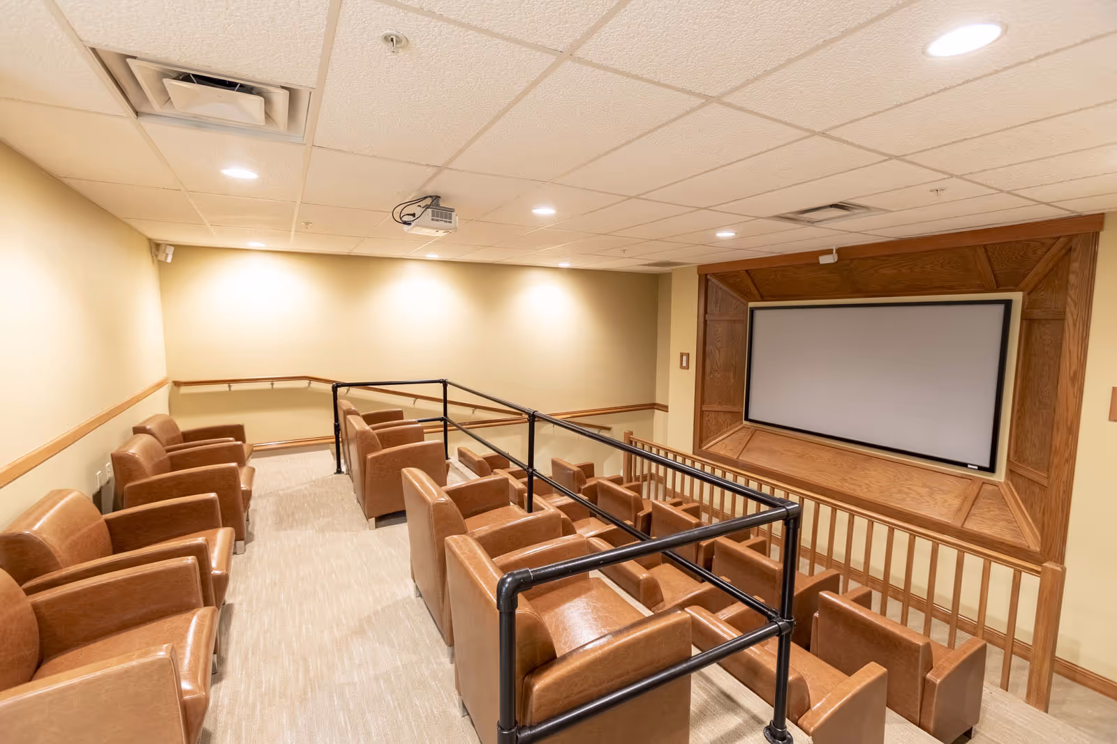 A small theater room with two rows of brown leather chairs facing a large blank projection screen mounted on a wooden frame. The room has beige walls, a carpeted floor, and a drop ceiling with recessed lighting and a ceiling-mounted projector.