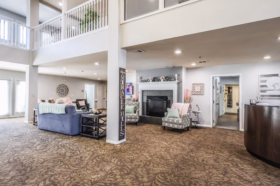 Spacious and well-lit senior living facility common area with patterned carpet, a blue sofa with a light green throw, two patterned armchairs with pastel cushions, a fireplace decorated with flowers, and a tall sign reading 'Welcome Miracle Hills'. The area has high ceilings with a balcony railing above and several doorways leading to other rooms.