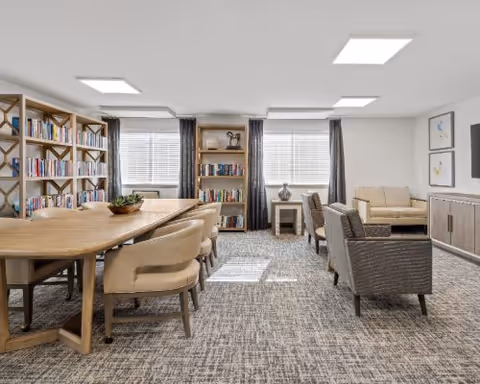 A bright and spacious senior living common area with a large wooden table surrounded by beige cushioned chairs on the left, bookshelves filled with books against the wall, two windows with blinds and dark curtains, a small side table with decorative items, and a seating area with a beige sofa and two gray armchairs on the right. The room has a patterned carpet and white ceiling with recessed lighting.
