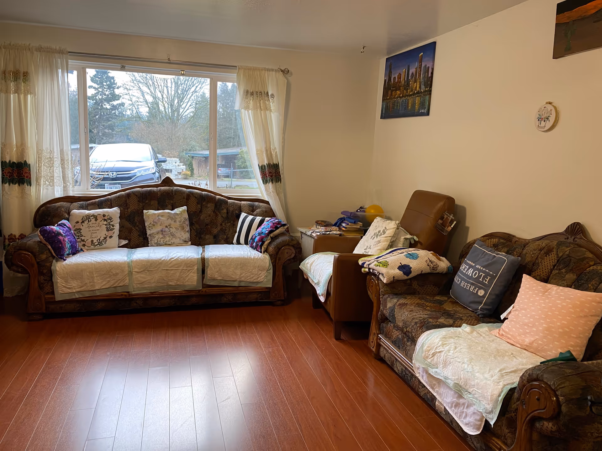 A cozy living room with two patterned sofas and a brown armchair. The sofas have various decorative pillows and white protective covers. A large window with sheer curtains lets in natural light and shows a parked car and trees outside. The room has wooden flooring and light-colored walls with a cityscape painting and small wall decorations.