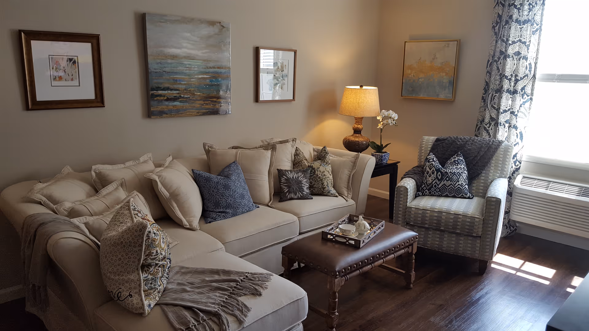 A cozy living room with a beige sectional sofa adorned with multiple decorative pillows and a throw blanket. There is a patterned armchair with pillows next to a window with patterned curtains. A wooden coffee table with a tray holding a teapot and cups sits in front of the sofa. The room has wooden flooring, a side table with a lamp and a small plant, and framed artwork on the walls.