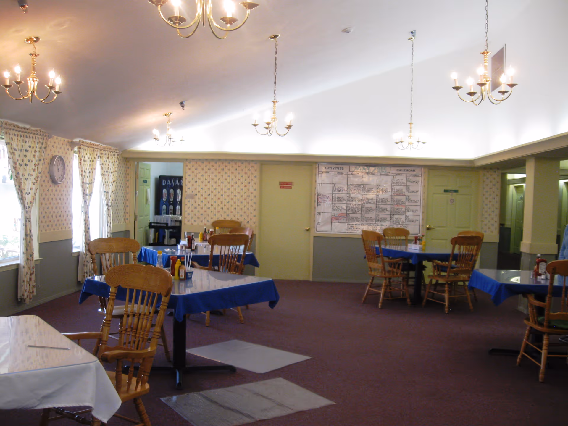 A dining room with several wooden chairs and tables covered with blue and white tablecloths. The room has patterned wallpaper, multiple chandeliers hanging from the ceiling, and windows with floral curtains. A large activities calendar is visible on the far wall, and a beverage vending machine is seen in the background.