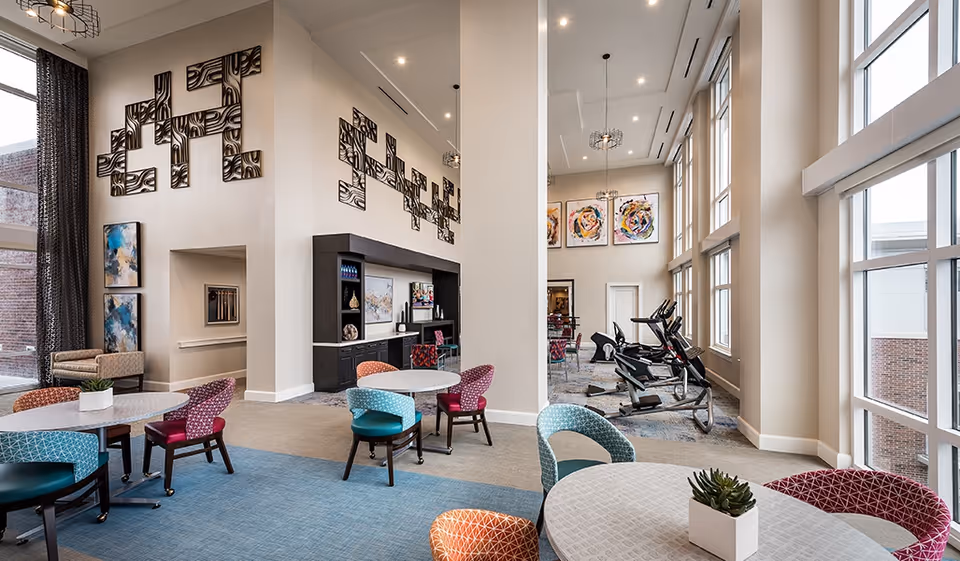 A bright and spacious common area in a senior living facility with high ceilings and large windows. The room features several round tables with colorful chairs, modern wall art, and a small exercise area with cardio equipment near the windows. The decor includes neutral walls, blue carpeting, and contemporary light fixtures.