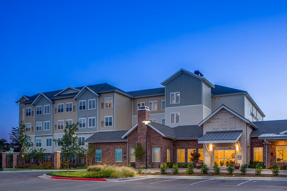 Front exterior of a multi-story senior living building with an illuminated entrance at dusk.