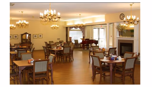 Spacious senior living dining room with wooden tables and chairs, chandeliers, a piano, and a fireplace.