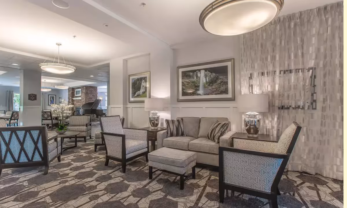 A well-lit living room area with a gray sofa, matching ottoman, and several upholstered armchairs arranged around small tables. The room features patterned carpet, modern lamps on side tables, framed nature photographs on the walls, and a grand piano in the background near a brick fireplace.