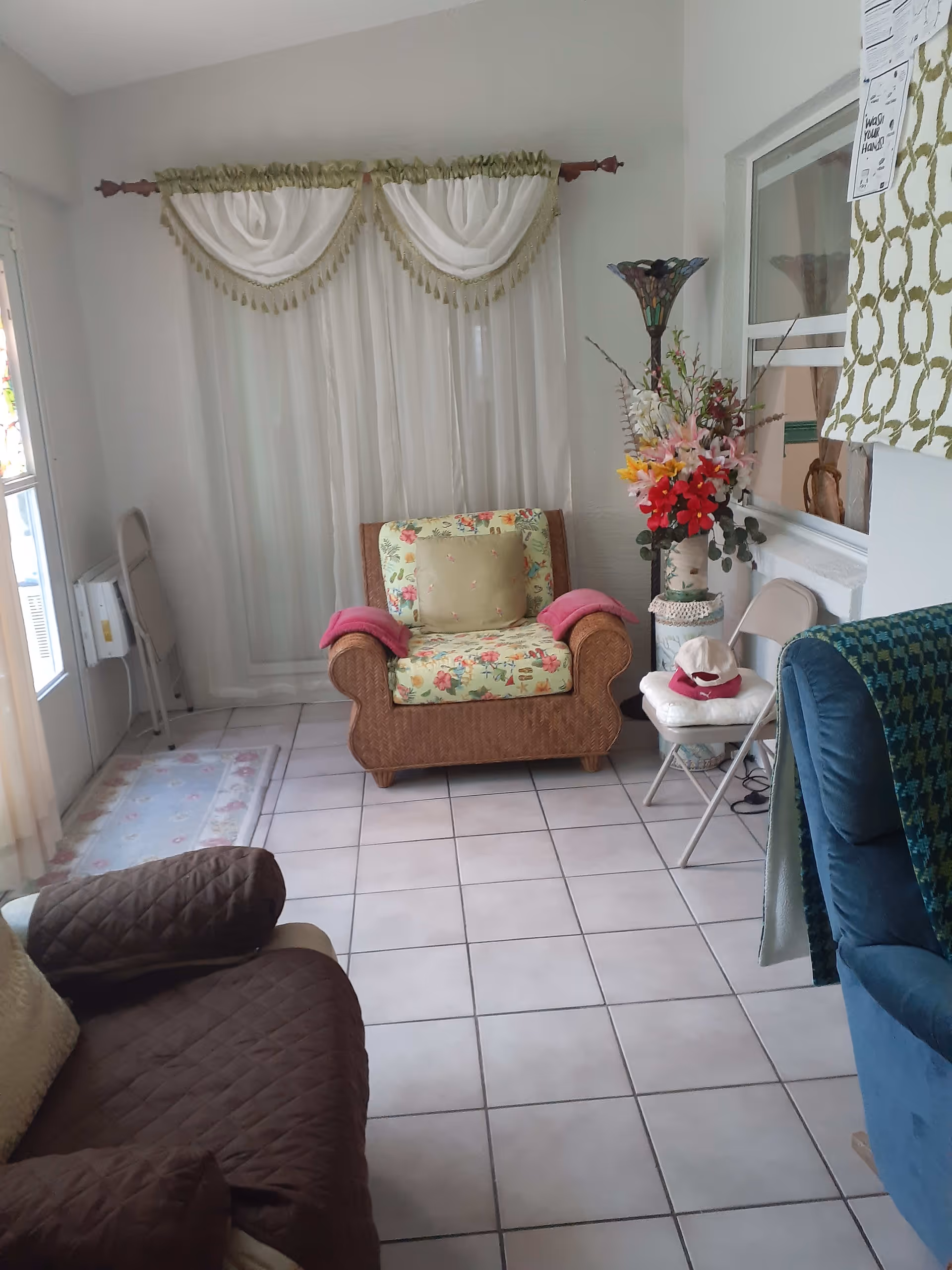 A cozy living room with tiled floor featuring a floral cushioned armchair with pink armrest covers, a brown quilted sofa, a blue armchair with a green patterned throw, a tall floor lamp with a stained glass shade, a vase with colorful artificial flowers, and white curtains with green and gold valances covering a window or door.