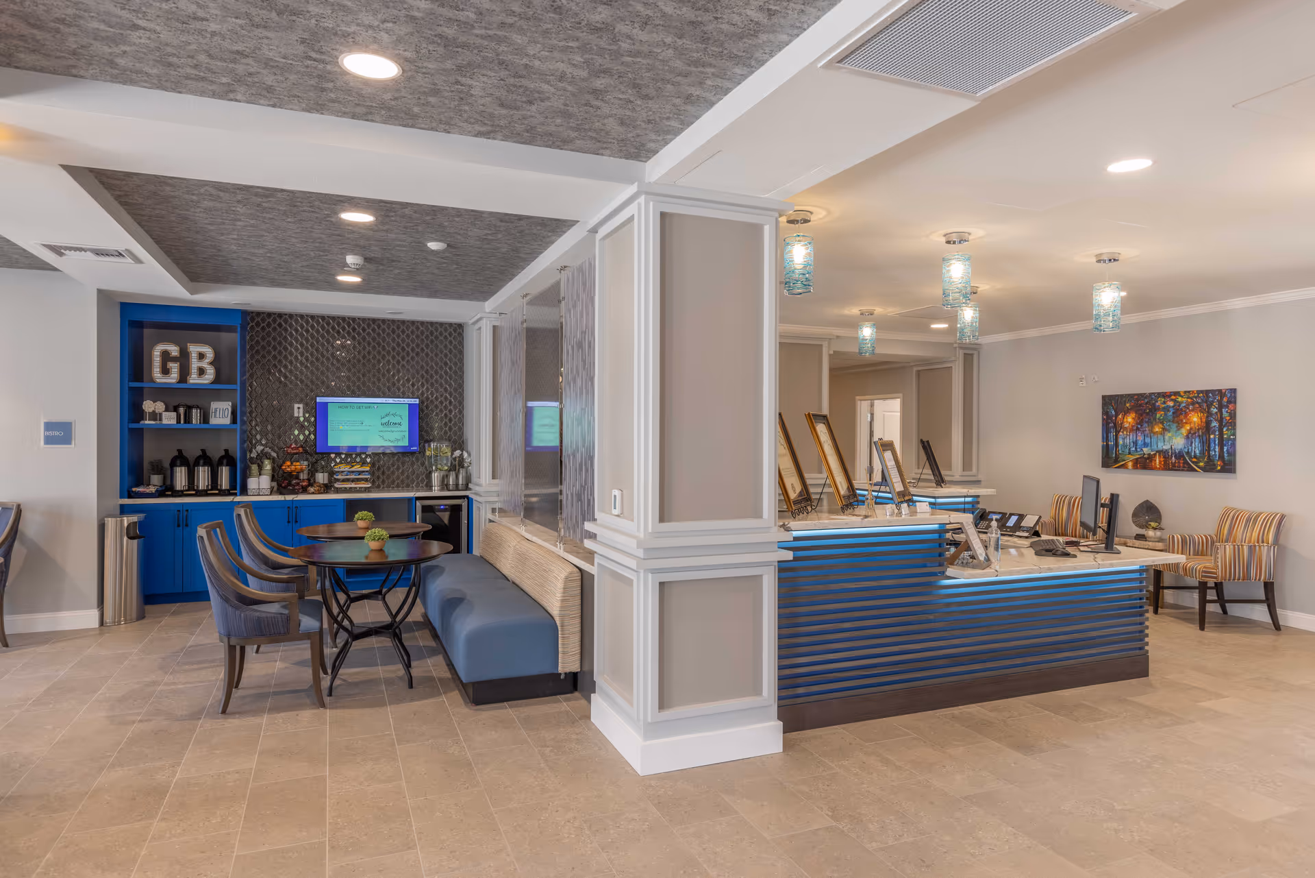 Interior view of a senior living facility lobby area with a reception desk on the right featuring computers and telephones, and a seating area on the left with tables, chairs, and a blue cushioned bench. Behind the seating area is a blue cabinet with coffee and snacks, a wall-mounted TV, and decorative items. The ceiling has recessed lighting and hanging pendant lights with blue glass shades.
