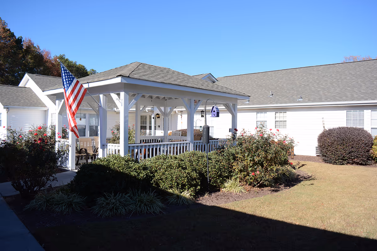 A white gazebo with an American flag in the landscaped courtyard of a single-story senior living building.