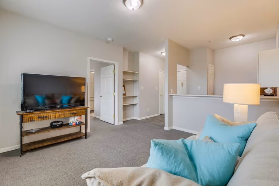 Bright living room with a sofa topped with teal pillows, a flat-screen TV on a wooden stand, and an open view into a hallway and kitchen counter.