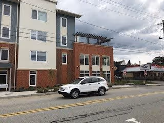 Exterior view of a multi-story senior living facility building with a white SUV parked in front on the street. The building features a combination of brick and light-colored siding with multiple windows and a pergola structure on the roof. Across the street, there is a small commercial building with signage.