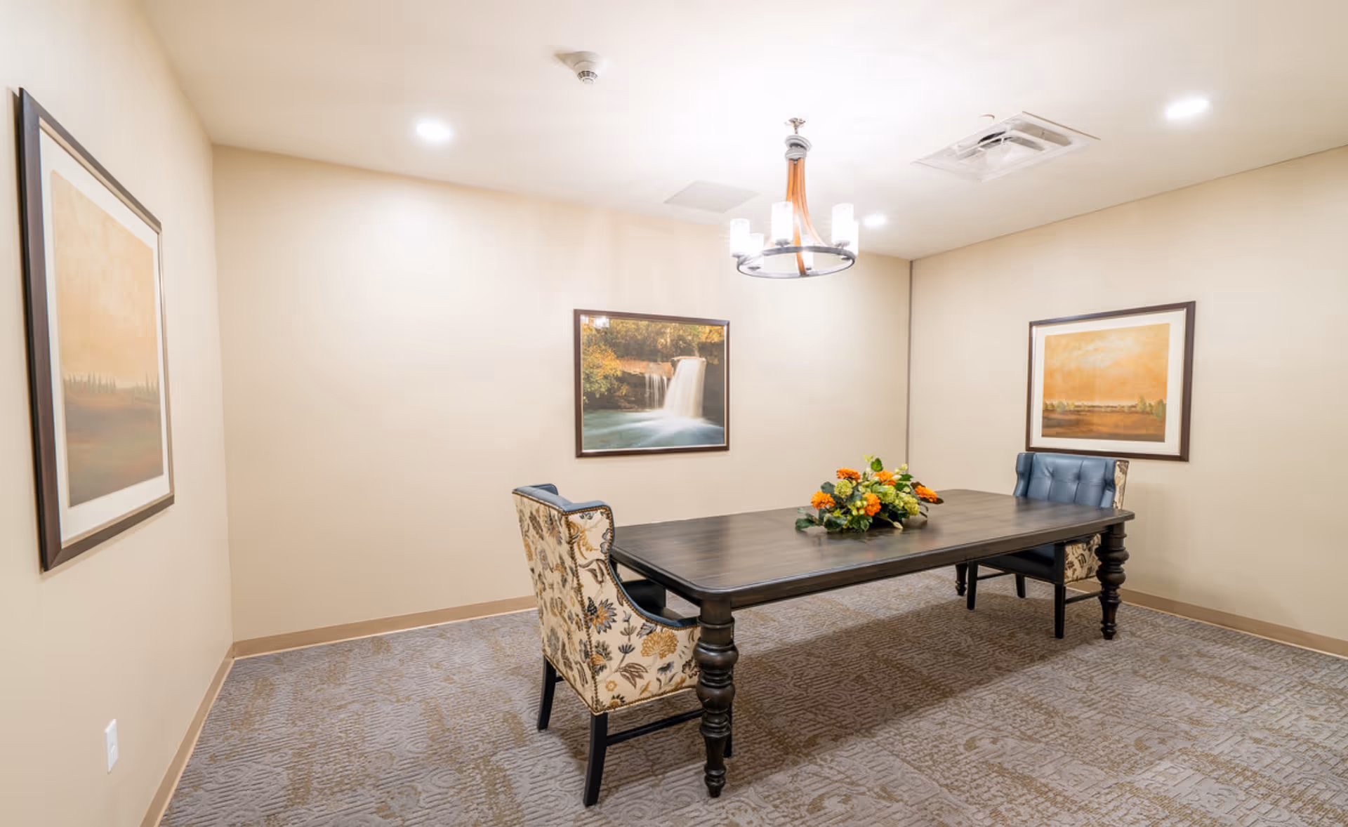 A small dining room with a dark wooden rectangular table and four chairs, two with floral upholstery and two with blue upholstery. A floral centerpiece is on the table. The room has beige walls with three framed landscape paintings and a modern chandelier hanging from the ceiling. The floor is carpeted with a patterned design.
