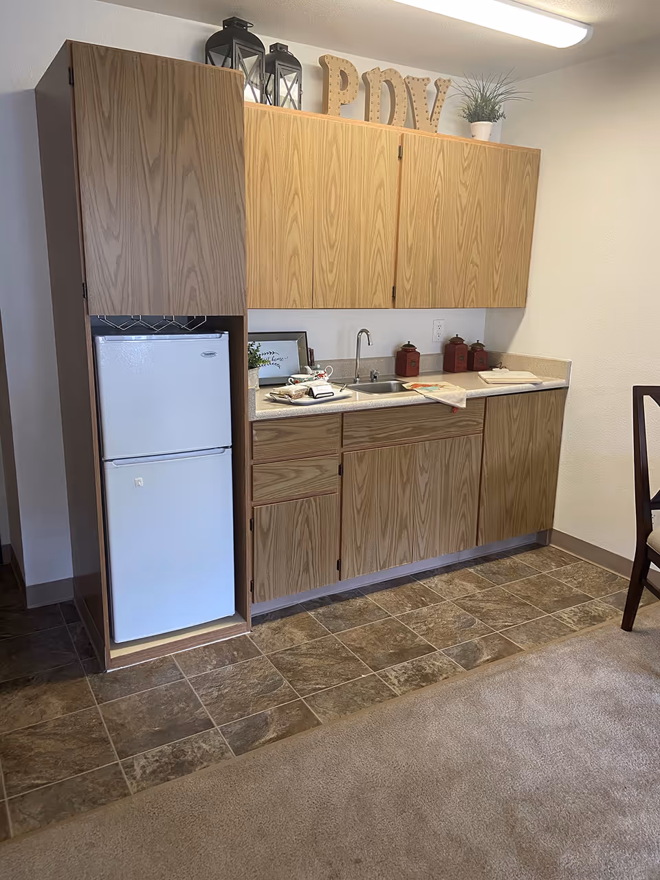 A small kitchenette area with wood cabinets, a white mini refrigerator, a stainless steel sink, and countertop with decorative items including three red canisters, a small plant, and a framed picture. The floor is tiled in front of the kitchenette and carpeted beyond it. A wooden chair is partially visible on the right side.