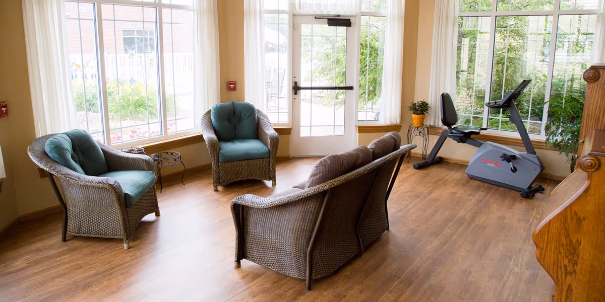 Sunlit lounge with wicker chairs, a sofa, and a stationary bike by large windows.