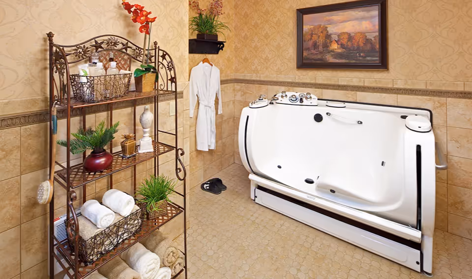 A bathroom with beige tiled walls and floor featuring a walk-in bathtub with safety handles and controls. A white bathrobe hangs on the wall next to the tub, with a pair of black slippers on the floor beneath it. To the left, there is a decorative metal shelving unit holding rolled towels, plants, and bath accessories. A framed landscape painting hangs on the wall above the bathtub.