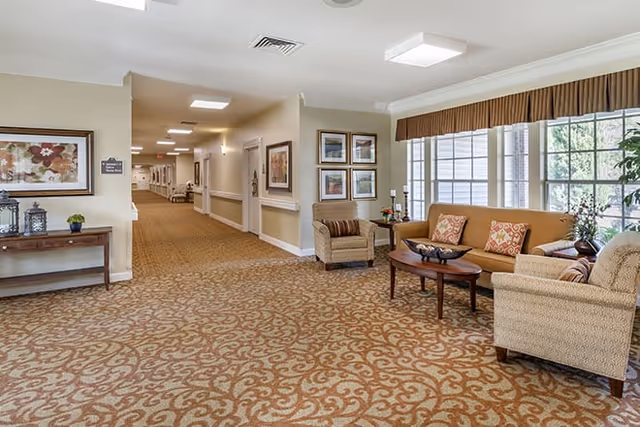 A spacious, well-lit hallway in a senior living facility with patterned carpet and beige walls. To the right, there is a seating area with a sofa, two armchairs, a wooden coffee table, and large windows with brown valances letting in natural light. The walls are decorated with framed artwork, and a wooden console table with decorative items is visible on the left side of the hallway.