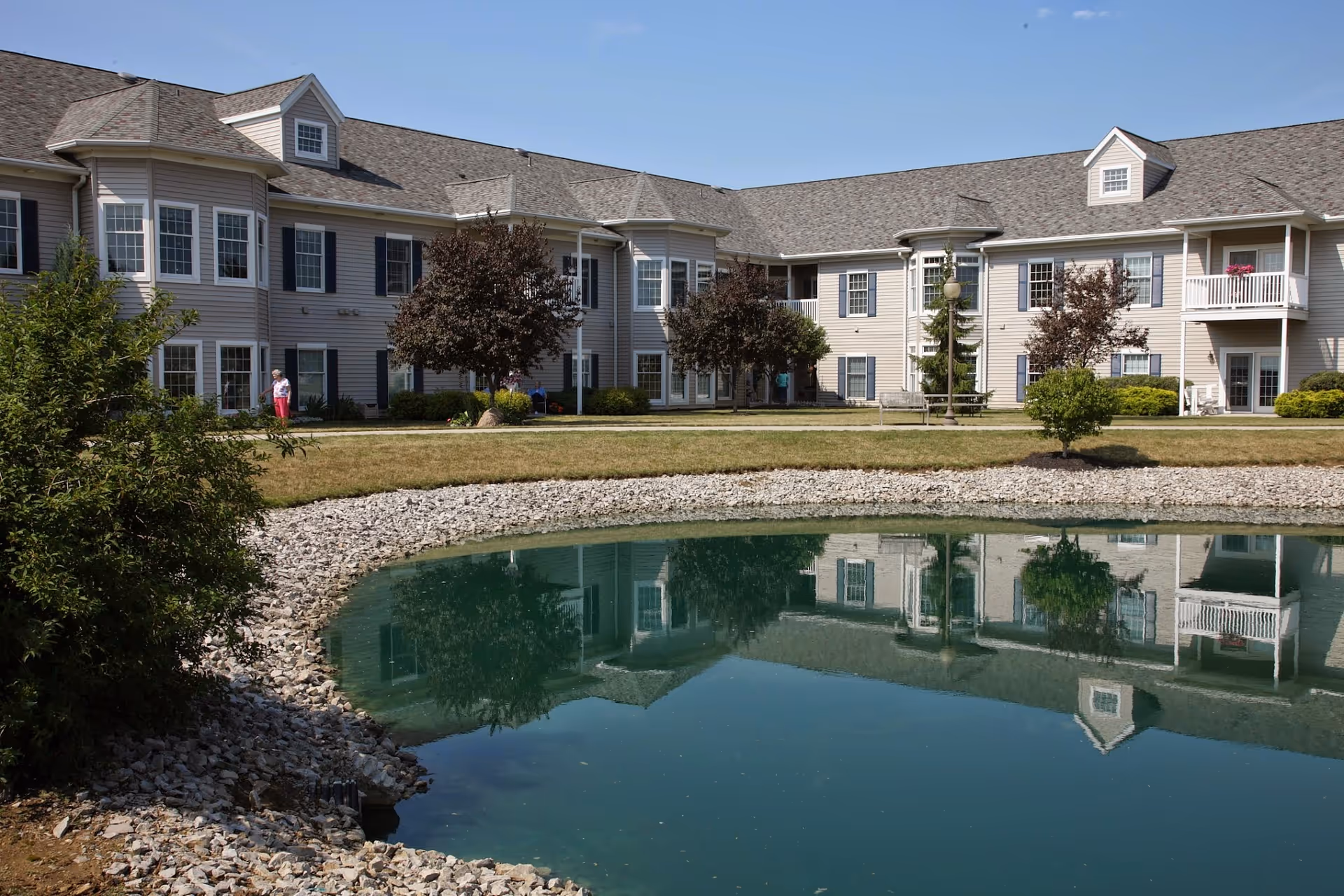 Exterior view of a senior living facility building with beige siding and multiple windows, reflected in a small pond surrounded by rocks. There are trees and bushes around the building and a person walking on the pathway near the building.