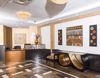 Reception area of Somerset Woods Rehabilitation & Nursing Center featuring a front desk with a chair, decorative wall art, two chairs with patterned cushions, a unique curved wooden bench, and a tiled floor with a geometric pattern. The ceiling has recessed lighting and two chandelier-style light fixtures.