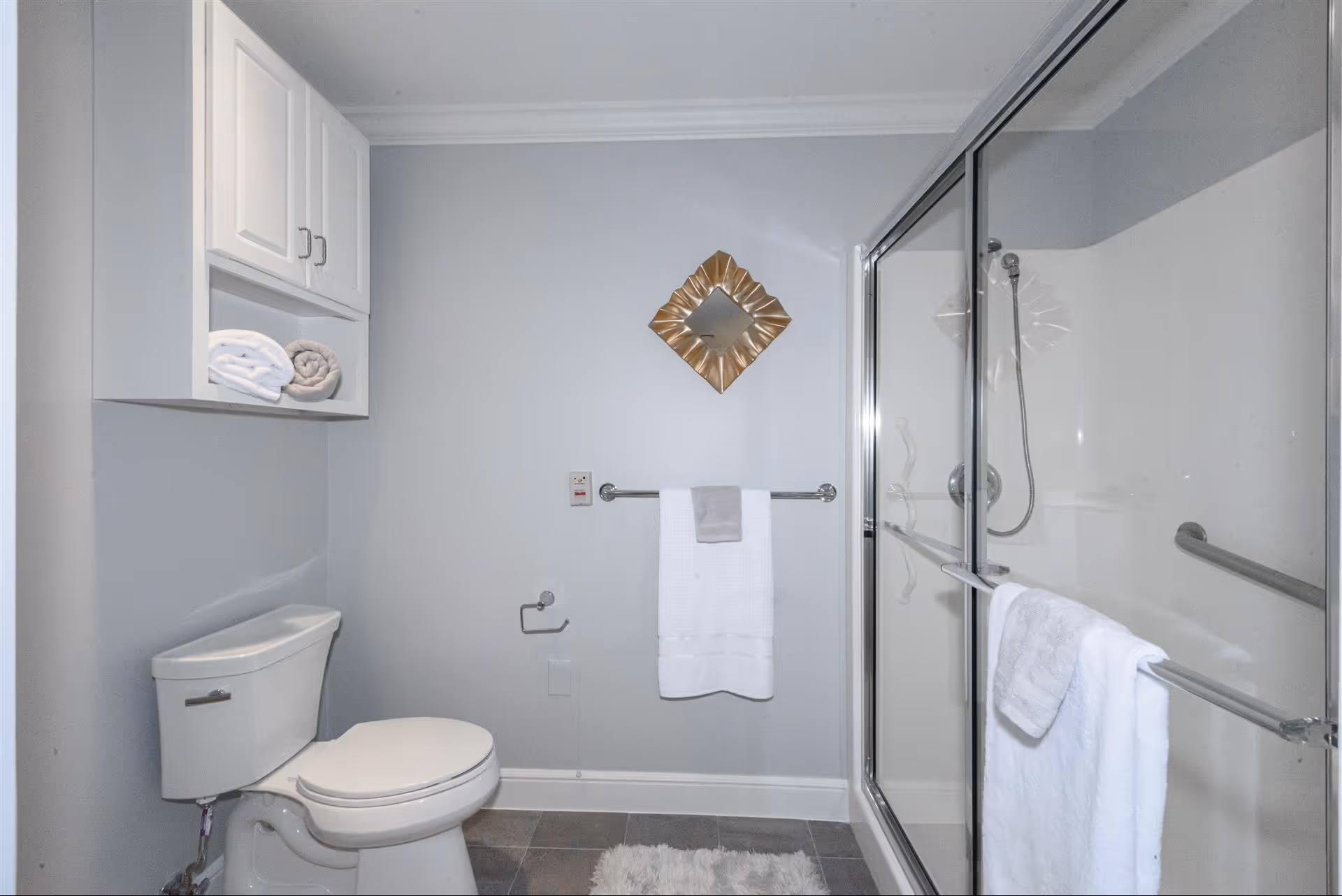 A clean bathroom with a white toilet, a glass-enclosed shower with a handheld showerhead, white towels hanging on a towel bar and the shower door, a small white cabinet mounted on the wall with rolled towels, a decorative square mirror with a gold frame on the wall, and a gray tiled floor with a white rug.