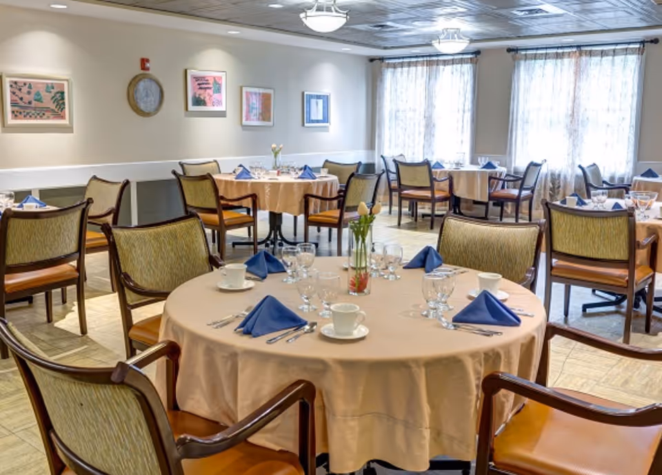 A dining room with round tables covered in beige tablecloths, each set with blue folded napkins, glassware, cups, and silverware. The room has several wooden chairs with cushioned seats and backs. The walls are decorated with framed artwork and a clock, and large windows with sheer curtains allow natural light to fill the space.