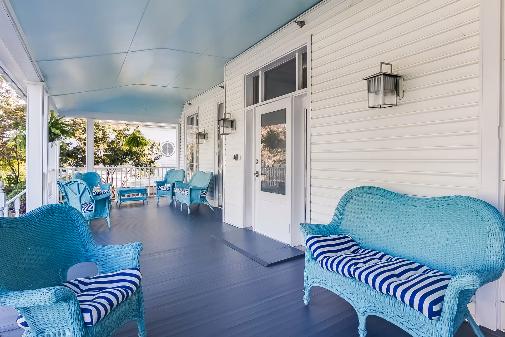Covered porch area with light blue wicker chairs and a loveseat, all with blue and white striped cushions. The porch has a blue floor and ceiling, white siding walls, and wall-mounted lantern-style lights. There are hanging plants and trees visible outside the porch railing.