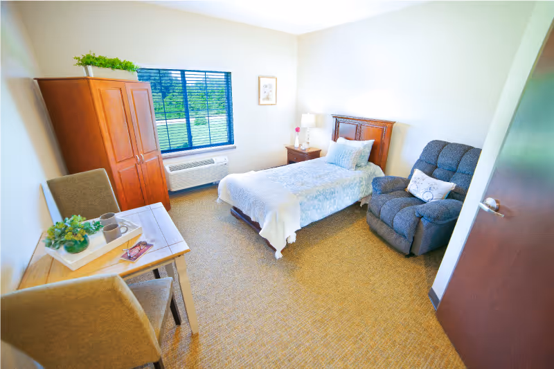 A bright and cozy bedroom in an assisted living facility featuring a single bed with light blue bedding, a wooden headboard, a nightstand with a lamp and flower vase, a comfortable gray recliner chair with a decorative pillow, a wooden wardrobe, a small table with two chairs, and a window with black blinds overlooking greenery outside.