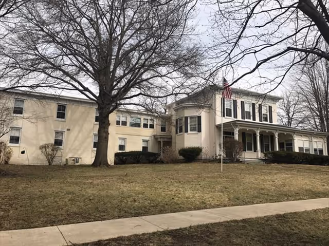 Exterior view of a large two-story beige building with black shutters and a covered porch. There is a flagpole with an American flag in front of the building, surrounded by leafless trees and a grassy lawn with a sidewalk in the foreground.