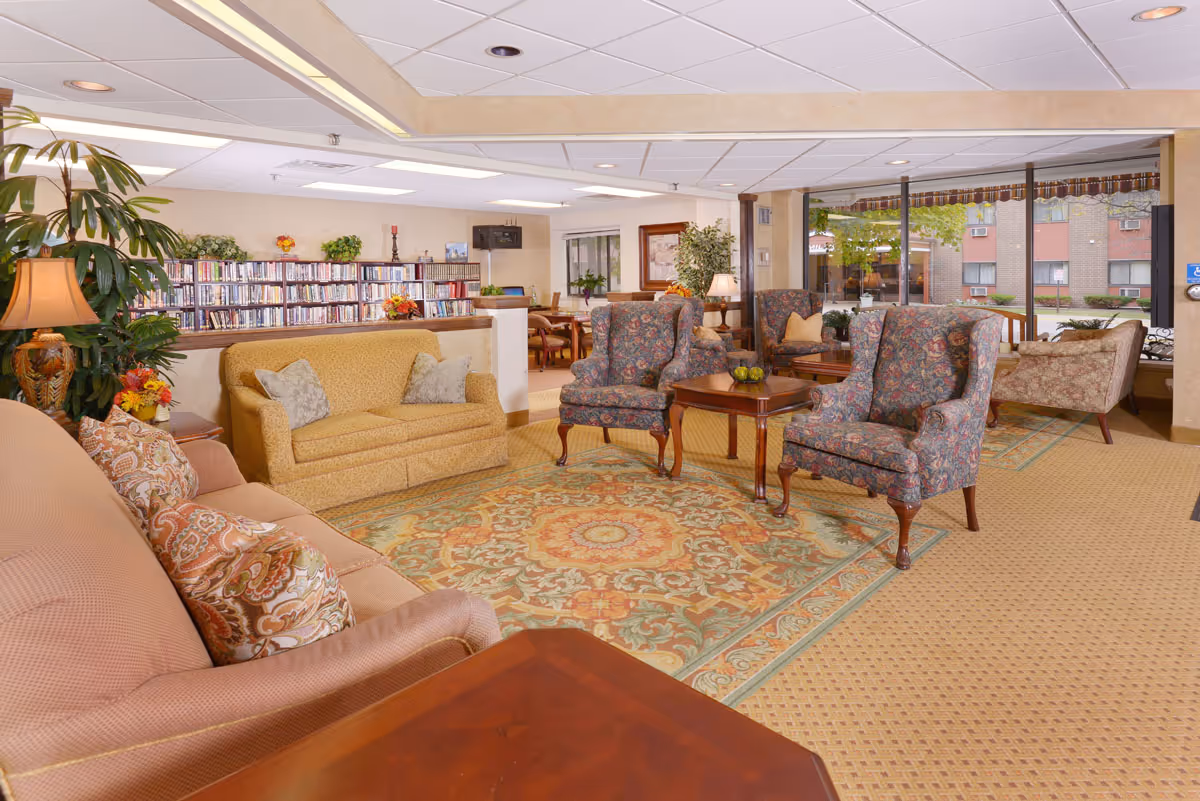 Well-lit senior living lounge with sofas, patterned armchairs, coffee tables, a large rug, and a bookshelf near windows.