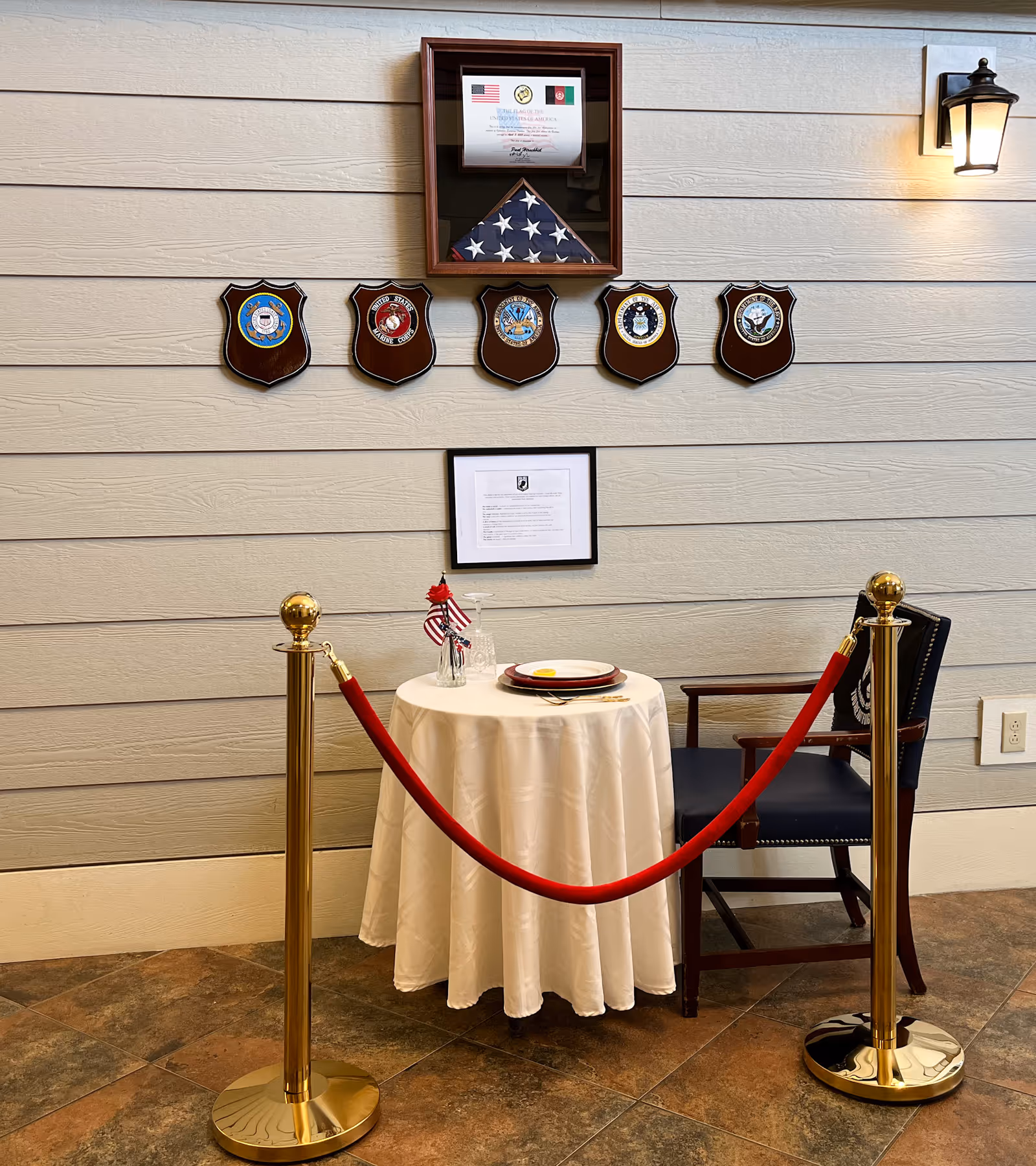 A small round table covered with a white tablecloth set with a plate, utensils, a glass, and a small vase holding a red flower and two small American flags. The table is roped off with a red velvet rope attached to two gold stanchions. Behind the table is a wooden chair with a dark cushion. On the wall above the table are five military service plaques, a folded American flag in a display case, and two framed documents. A wall-mounted lantern light is visible on the upper right side.