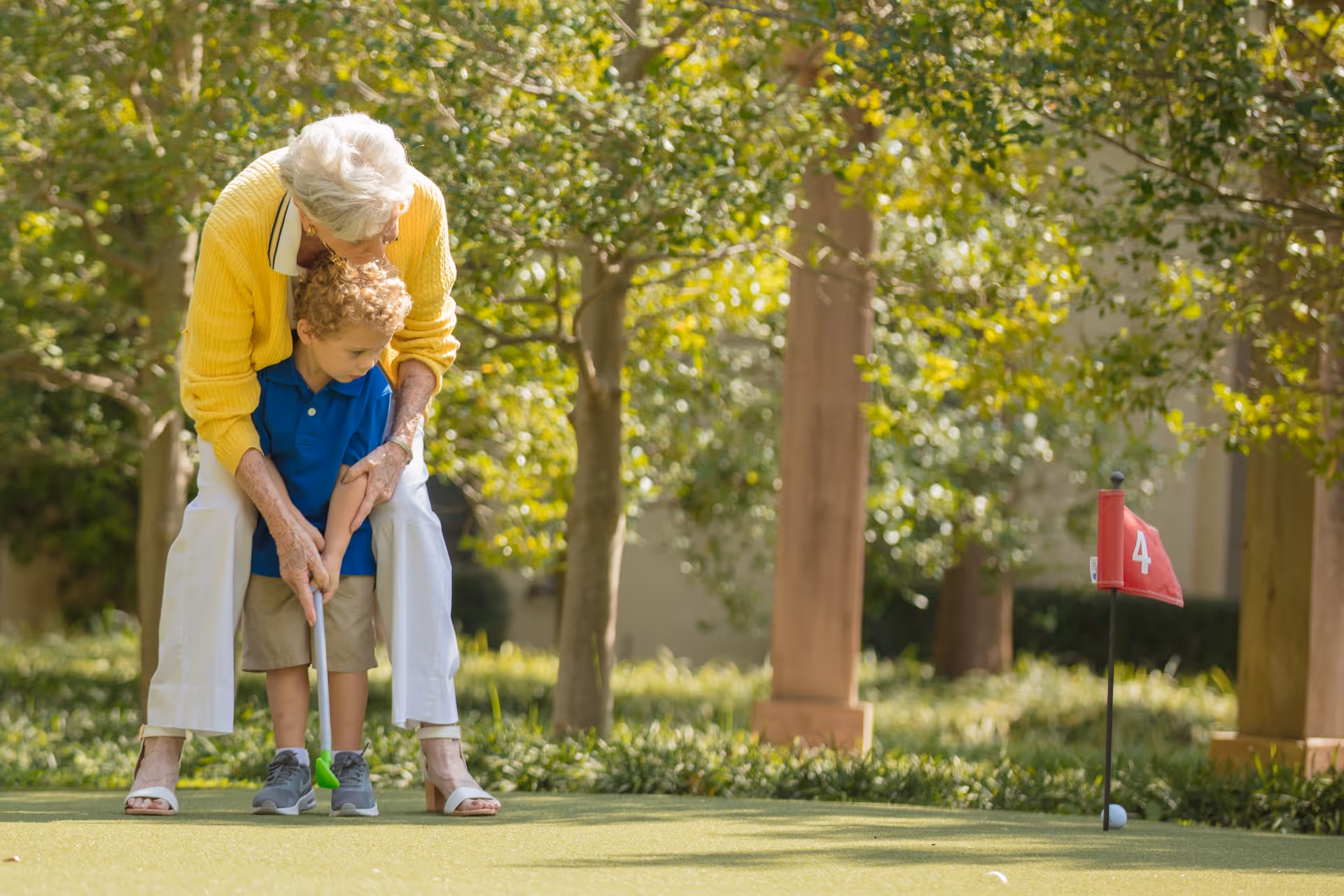 An elderly woman in a yellow sweater is helping a young boy in a blue shirt and beige shorts to putt a golf ball on a putting green with a red flag marked number 4, surrounded by trees and greenery.