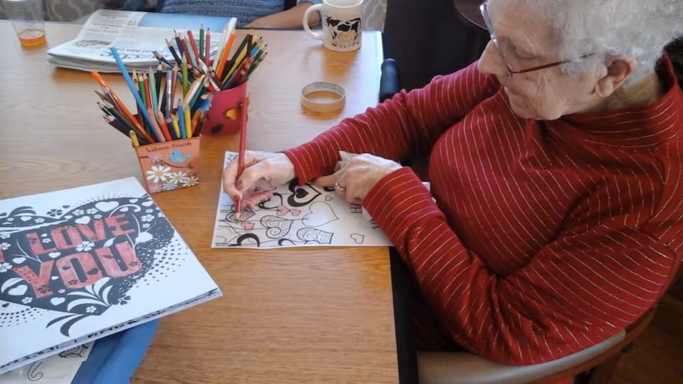 An elderly woman wearing glasses and a red striped sweater is sitting at a wooden table, coloring a heart-themed design on a piece of paper with colored pencils. On the table, there is a container filled with various colored pencils and a coloring book with a cover that says 'LOVE YOU' in large letters.