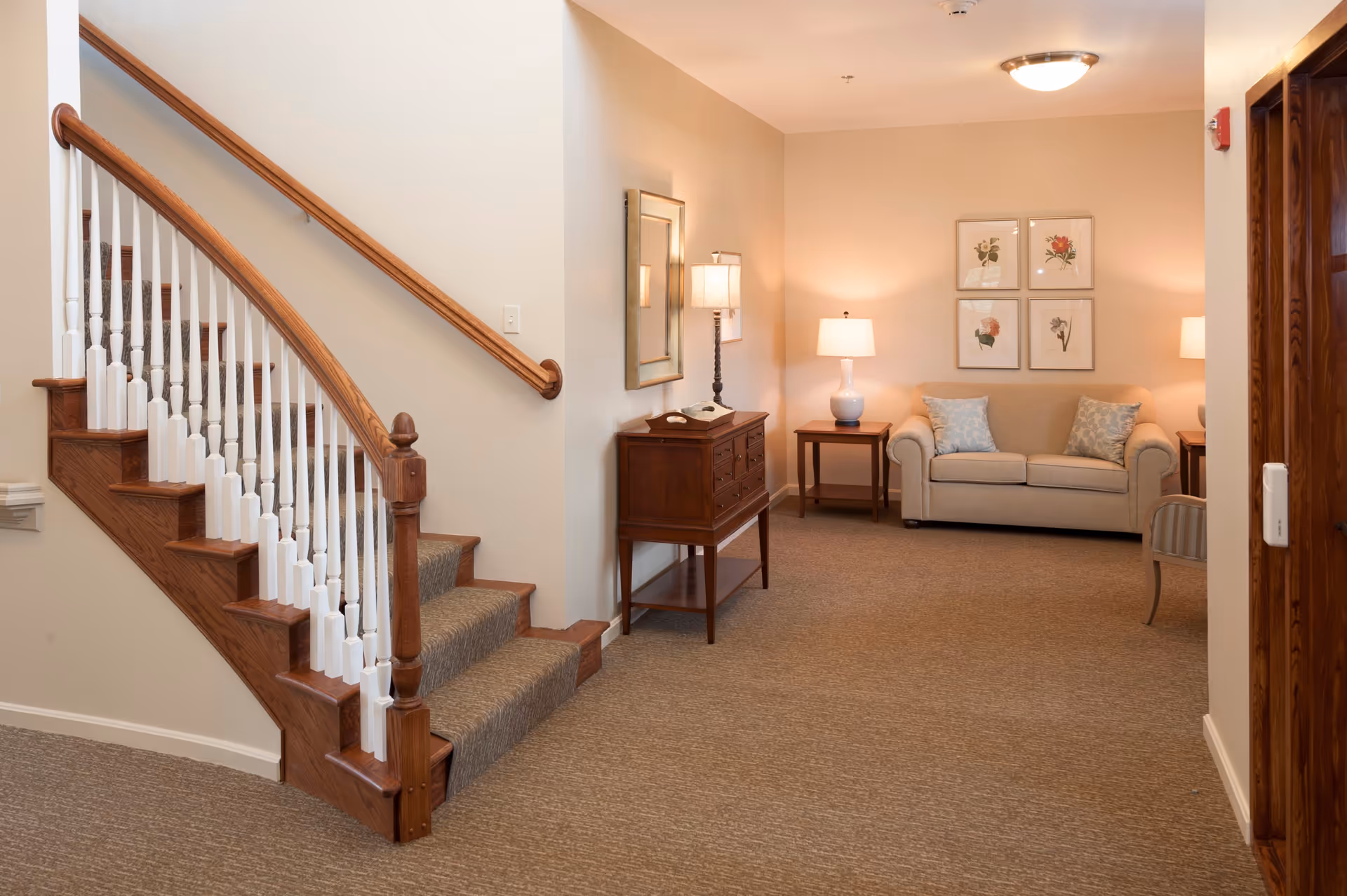 Interior view of a senior living facility showing a carpeted staircase with wooden handrails on the left and a cozy sitting area on the right. The sitting area includes a beige loveseat with two patterned pillows, two wooden side tables each with a lamp, a wooden console table with a lamp and a mirror above it, and four framed botanical prints on the wall. The space is softly lit with warm lighting.