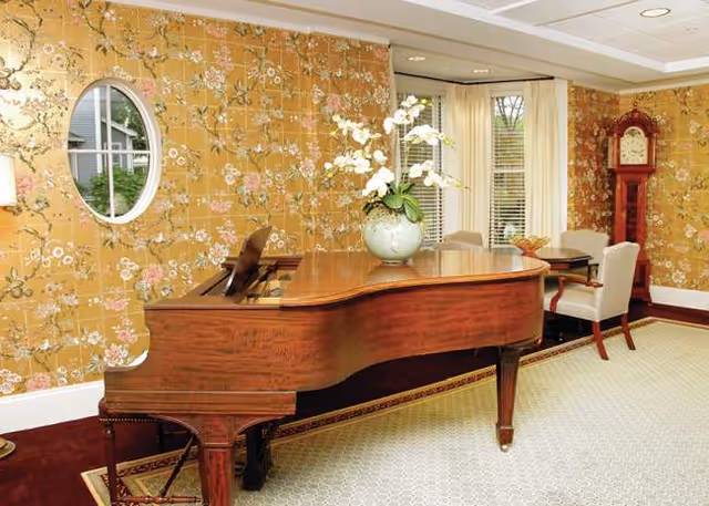 A grand piano sits in a floral lounge with chairs, orchid centerpiece, and a grandfather clock.