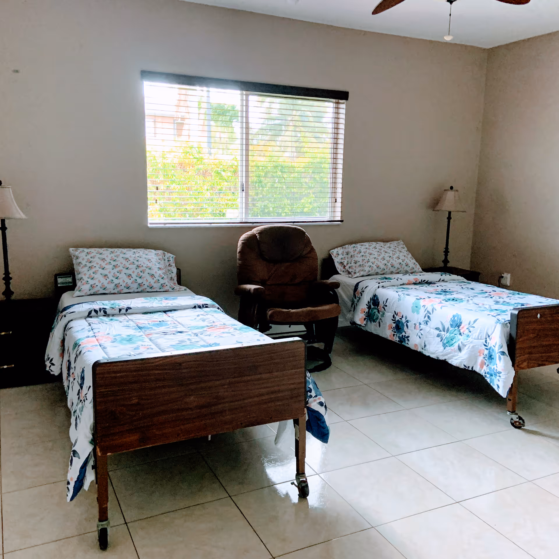 A simple bedroom with two single beds on wheels, each with floral bedding and a pillow. Between the beds is a brown cushioned armchair. There are two bedside tables, each with a lamp. A window with blinds is centered on the wall behind the beds, letting in natural light and showing greenery outside. The floor is tiled and the walls are plain.