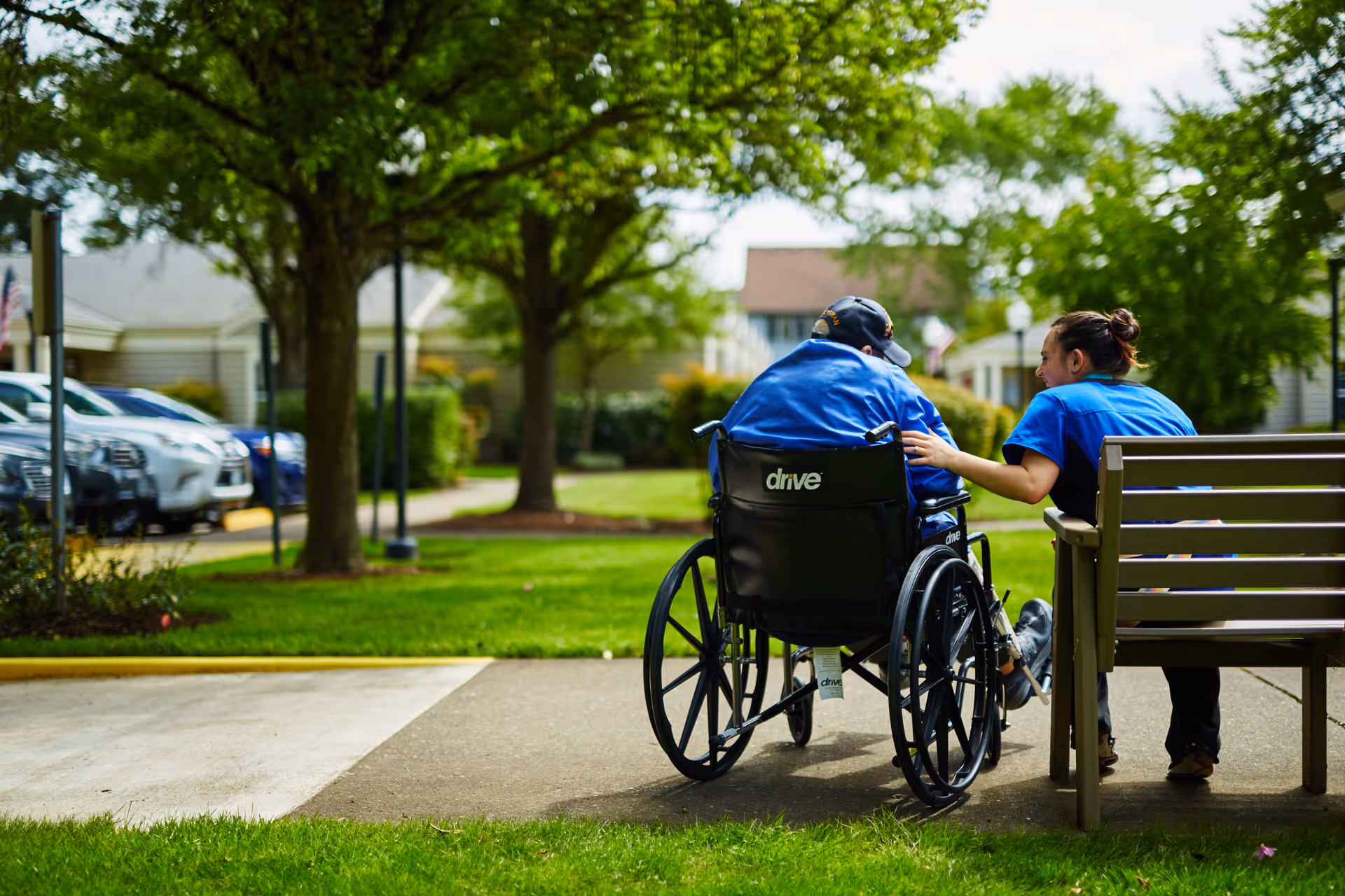 A person in a wheelchair is outdoors on a paved path next to a bench where a caregiver or companion is sitting, gently touching the person's shoulder. They are surrounded by green grass, trees, and residential buildings in the background under a partly cloudy sky.