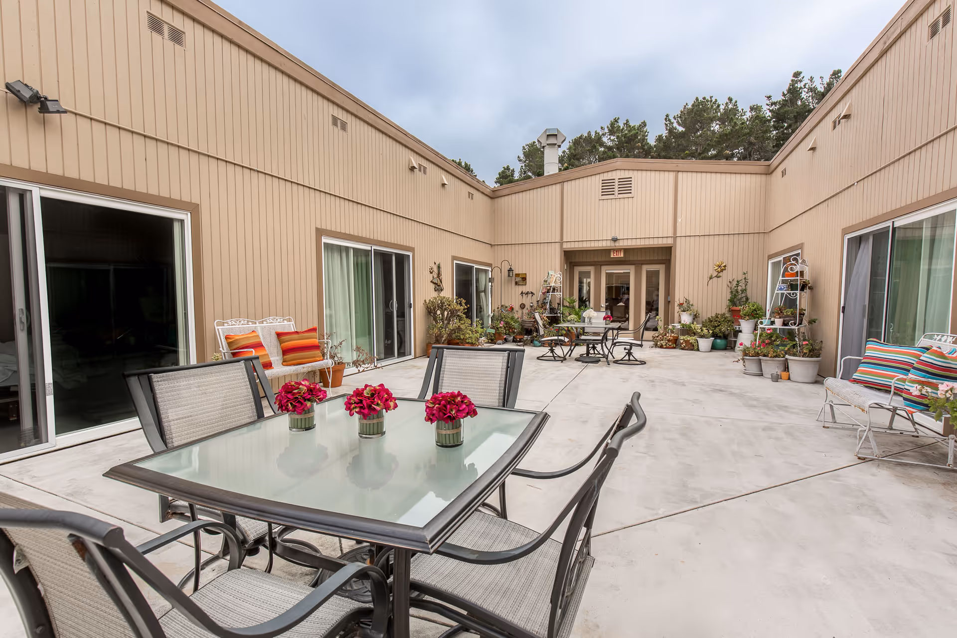 Outdoor courtyard area with a glass-top table and four chairs in the foreground, decorated with three small pots of red flowers. The courtyard is surrounded by beige walls with sliding glass doors and windows. There are several potted plants and outdoor seating arrangements with cushions along the walls. The sky is overcast.