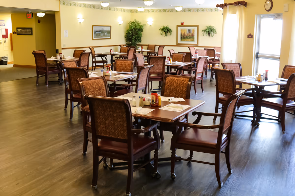A dining room in a senior living facility with multiple wooden tables and chairs arranged neatly. Each table is set with placemats, napkins, and condiments including ketchup, mustard, salt, and pepper. The room has yellow walls adorned with framed pictures and green plants. There is a clock above a door with curtains letting in natural light.