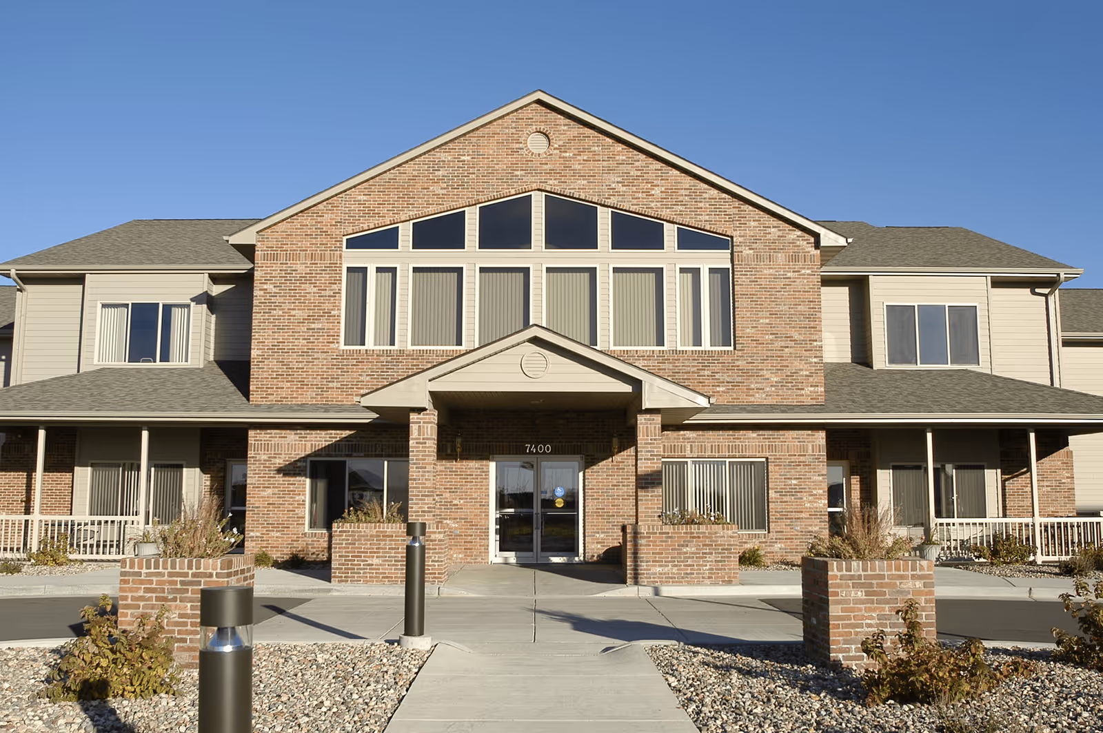 Front exterior view of a two-story senior living facility building with a brick facade and large windows under a clear blue sky. The entrance has a covered porch with double glass doors and the number 7400 above it. There are landscaped areas with rocks and plants along the walkway leading to the entrance.