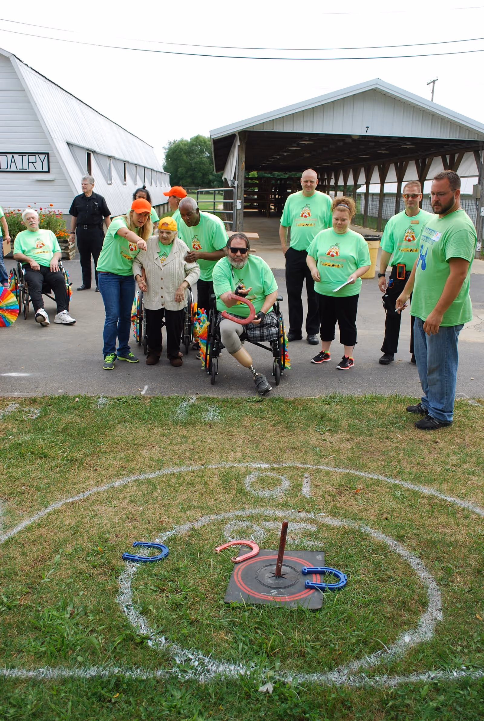A group of people, some in wheelchairs and others standing, are playing a ring toss game outdoors near a barn-like building with a sign that says 'DAIRY'. Most participants are wearing matching bright green t-shirts, and one person is in a wheelchair tossing a ring towards the target on the grass.