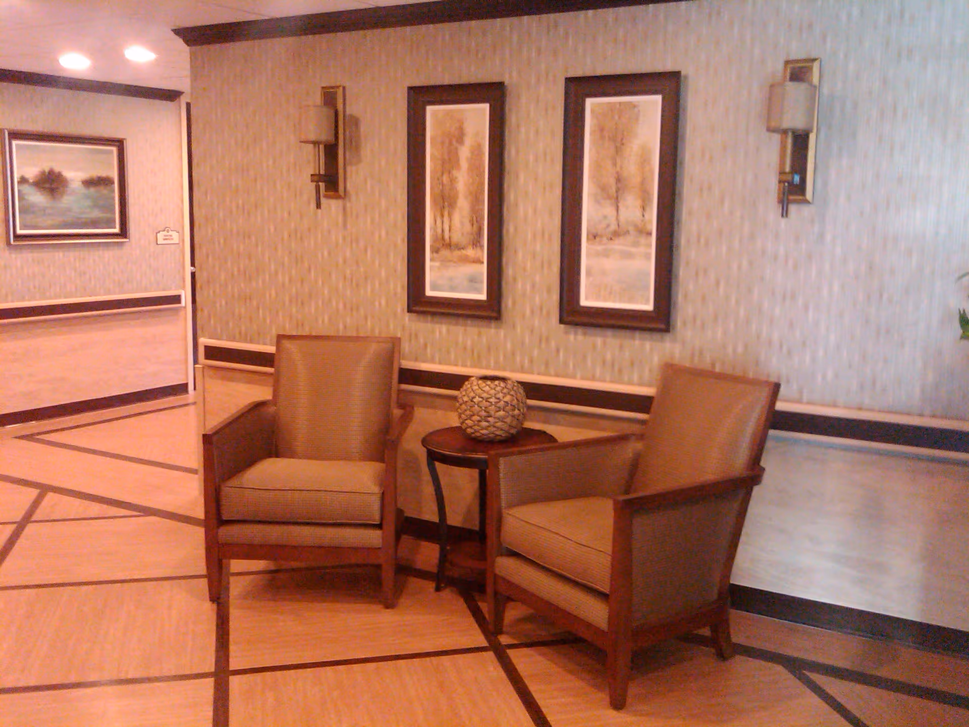 A seating area with two brown upholstered armchairs and a small round wooden table between them, holding a decorative spherical object. The wall behind features two framed vertical landscape paintings and two wall-mounted light fixtures. The floor has a patterned design with wood tones, and there is another framed painting visible in the adjacent hallway.