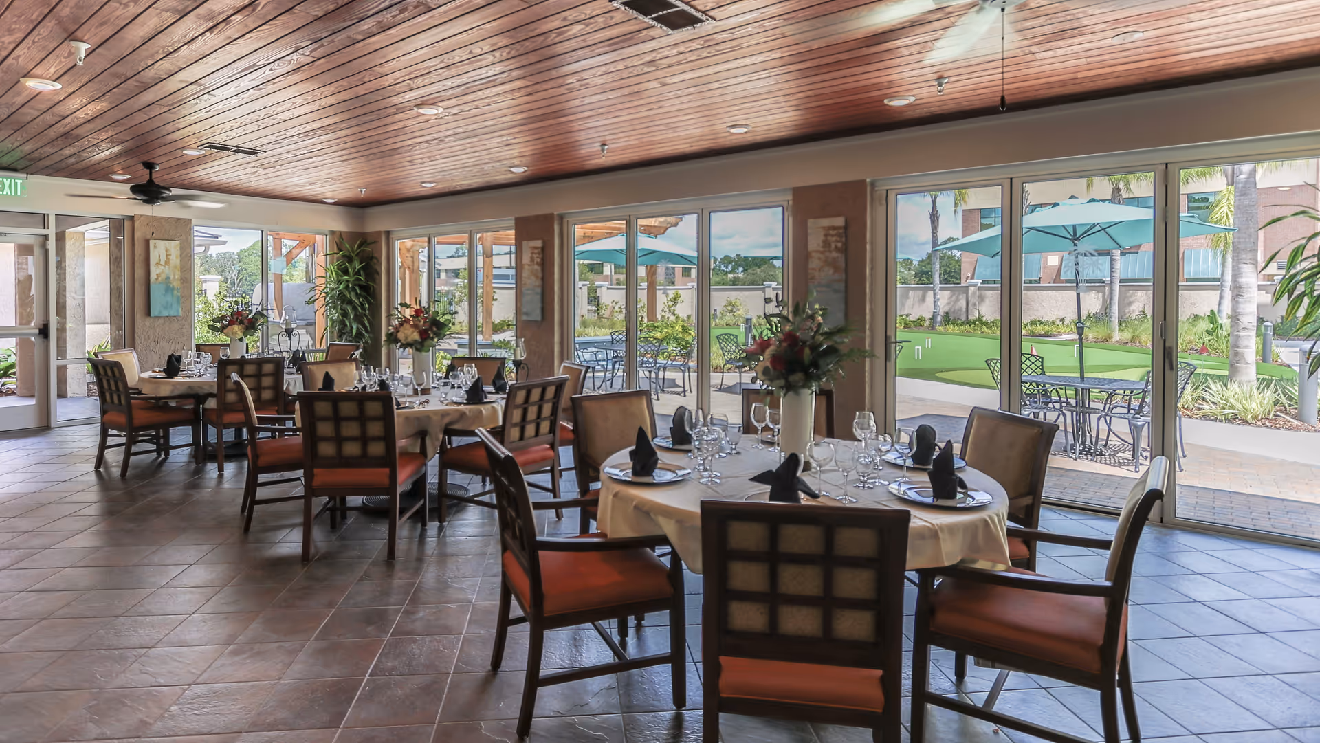 A dining room with round tables set with white tablecloths, black napkins, glassware, and floral centerpieces. The room has large windows and glass doors that open to an outdoor patio area with tables, chairs, and umbrellas. The ceiling is wooden with recessed lighting and ceiling fans.