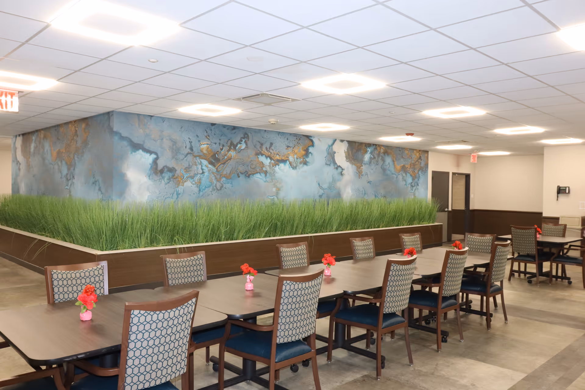 A spacious dining area with multiple wooden tables and patterned chairs arranged neatly. Each table has small red flower centerpieces. A large planter box with tall green grass separates the dining space from a wall featuring an abstract blue and gold mural. The ceiling has recessed lighting and there are exit signs visible in the background.