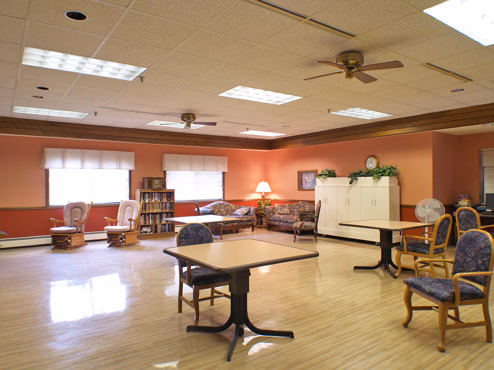 A spacious room with wooden flooring and peach-colored walls featuring several tables with chairs, two cushioned rocking chairs near windows with white valances, a bookshelf, floral-patterned sofas, a lamp on a side table, a white cabinet with plants on top, and ceiling fans with fluorescent lights.