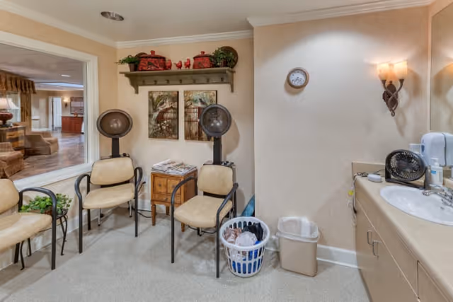 Interior view of a senior living facility room with three beige chairs arranged near two vintage hair dryers. A small wooden side table with magazines is placed between the chairs. On the right side, there is a countertop with a sink, a small fan, and a soap dispenser. A laundry basket and a trash bin are on the floor next to the counter. The walls are decorated with two framed pictures, a small clock, and a wall-mounted light fixture. A window on the left side shows a glimpse of another room with seating and wooden flooring.