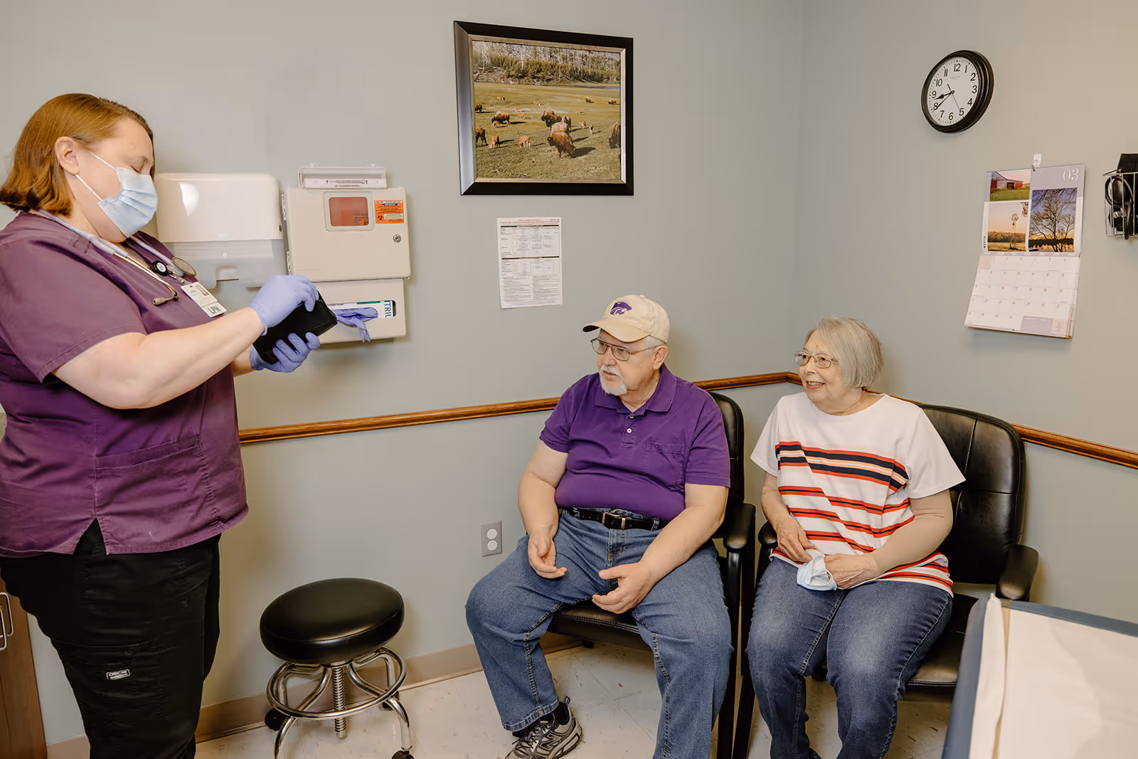 A healthcare worker wearing a mask and purple scrubs is putting on gloves in a medical examination room. Two elderly patients, a man in a purple shirt and cap and a woman in a white shirt with red stripes, are seated on black chairs against the wall. The room has light gray walls, a clock, a calendar, a framed picture of bison, and medical equipment on the walls.