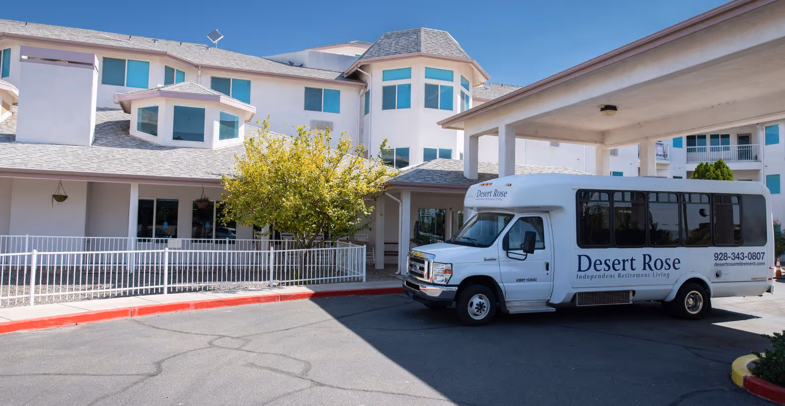 Exterior view of Desert Rose Retirement Community building with a white shuttle bus parked under a covered entrance. The bus has the Desert Rose logo and contact information on its side. The building is multi-story with multiple windows and a small tree near the entrance.